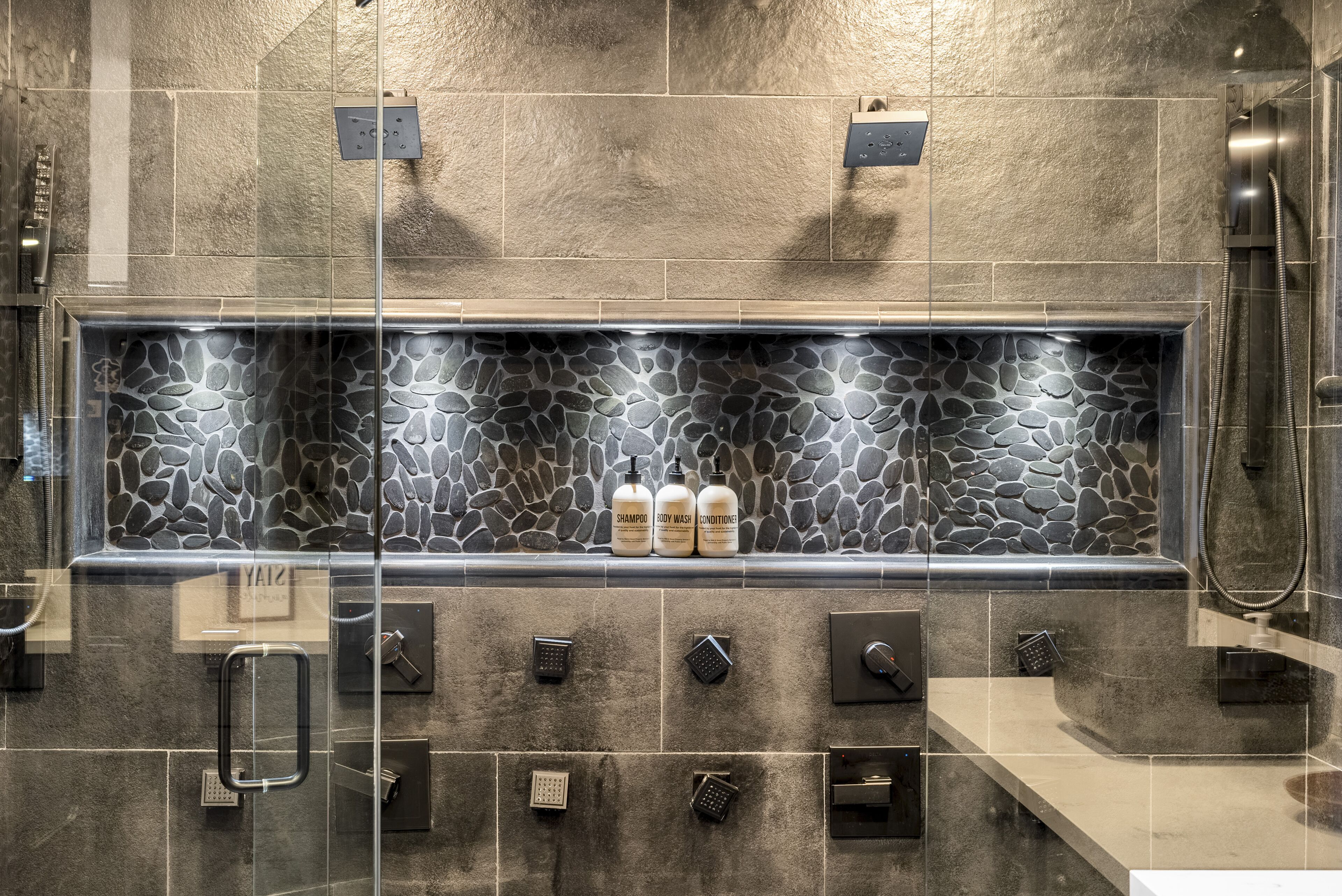 A modern bathroom with a stone mosaic backsplash, multiple showerheads, and a glass-enclosed shower area.