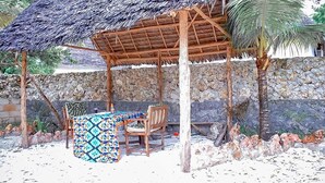 A covered outdoor seating area with a table and chairs, a thatched roof, and a stone wall in the background.