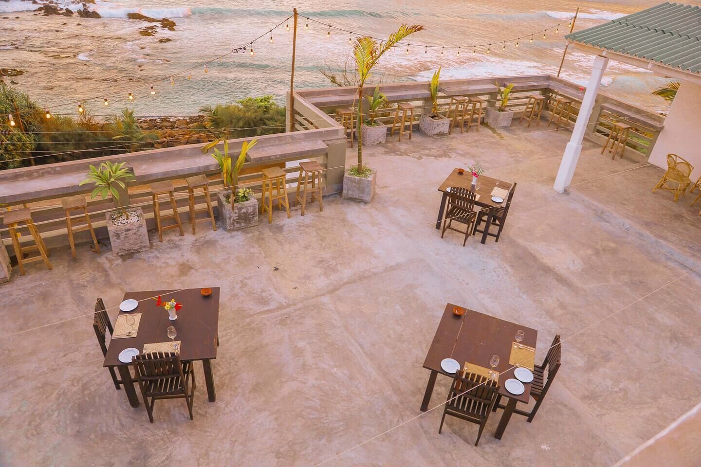 An outdoor patio with four tables and chairs, surrounded by potted plants and string lights.