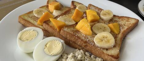 Une assiette de petit-déjeuner avec des toasts, des bananes, des morceaux de mangue et des œufs, une théière blanche et une tasse de café sur une table.