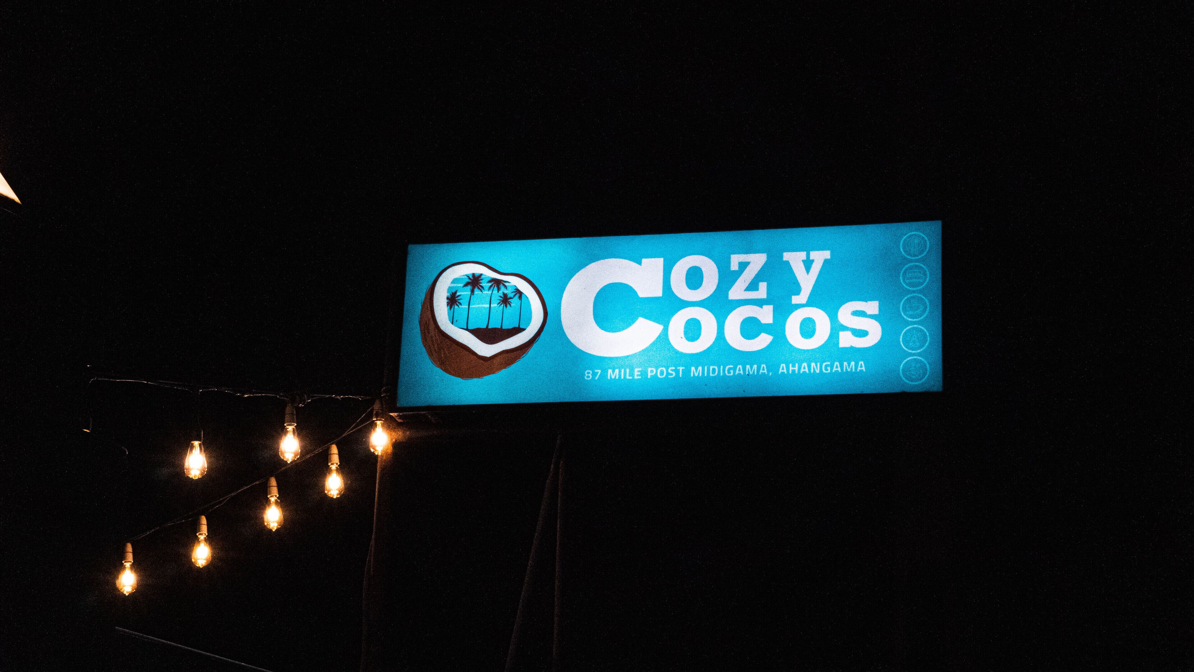 Letrero de 'Cozy Cocos' con logo de escena de playa, ubicado a 87 millas después de midigama, ahangama.
