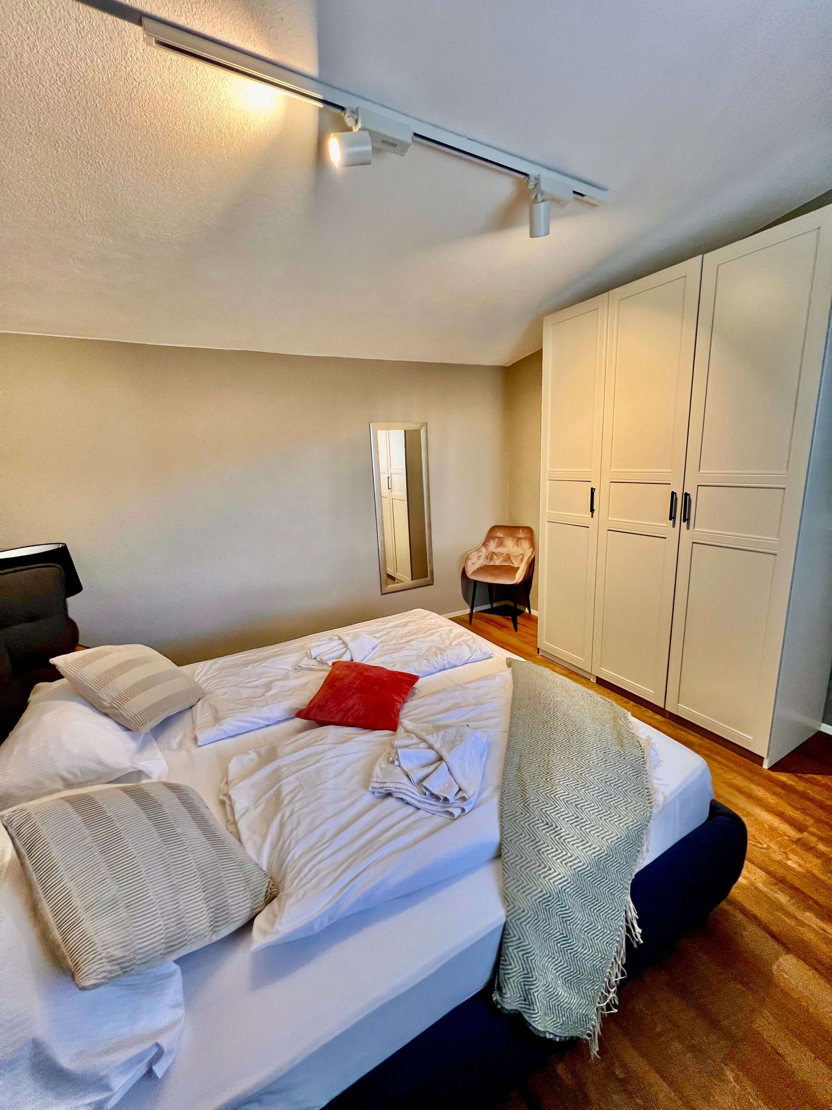 A neatly made bed with pillows and a blanket, a wardrobe, a chair, and a mirror on the wall.