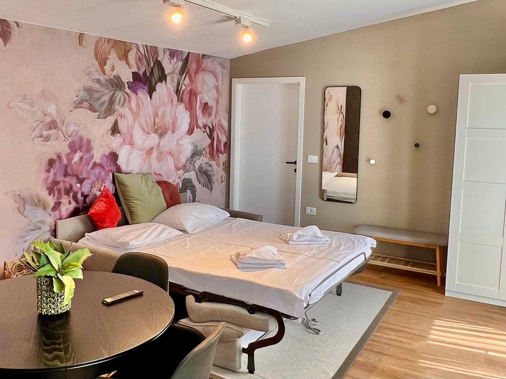 A hotel room with a bed, a round table with chairs, a large floral wall mural, a mirror, and a wardrobe.