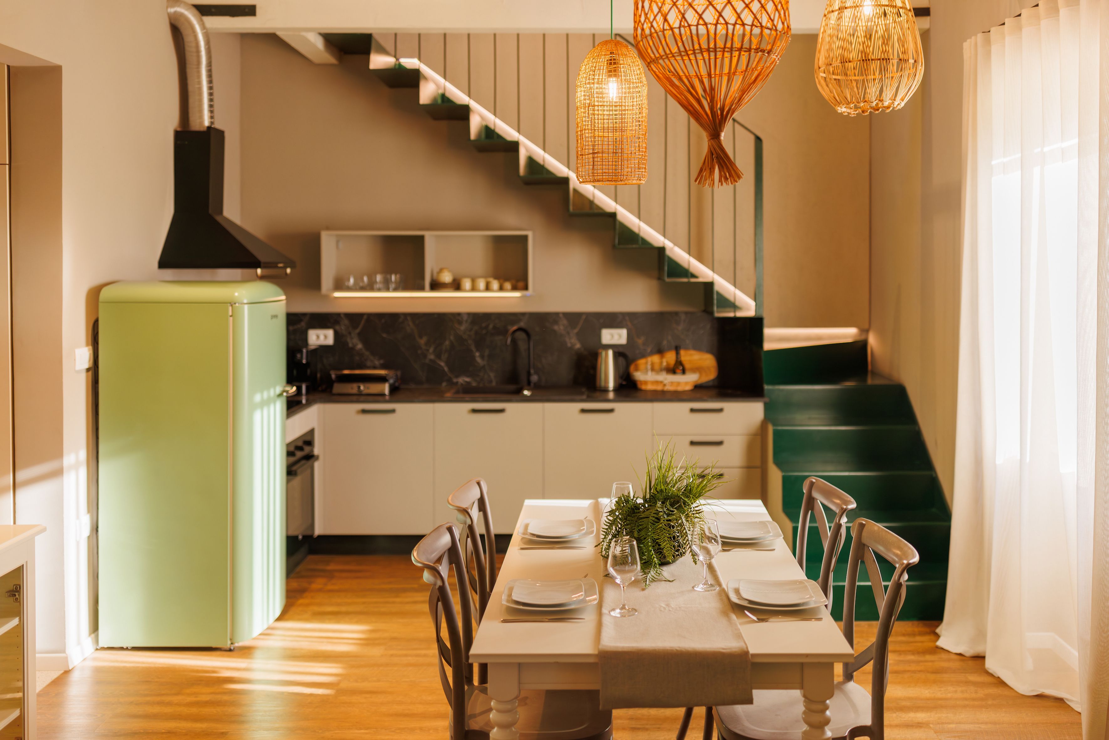 A modern kitchen with a green refrigerator, a dining table set for two, and a staircase leading to an upper level.
