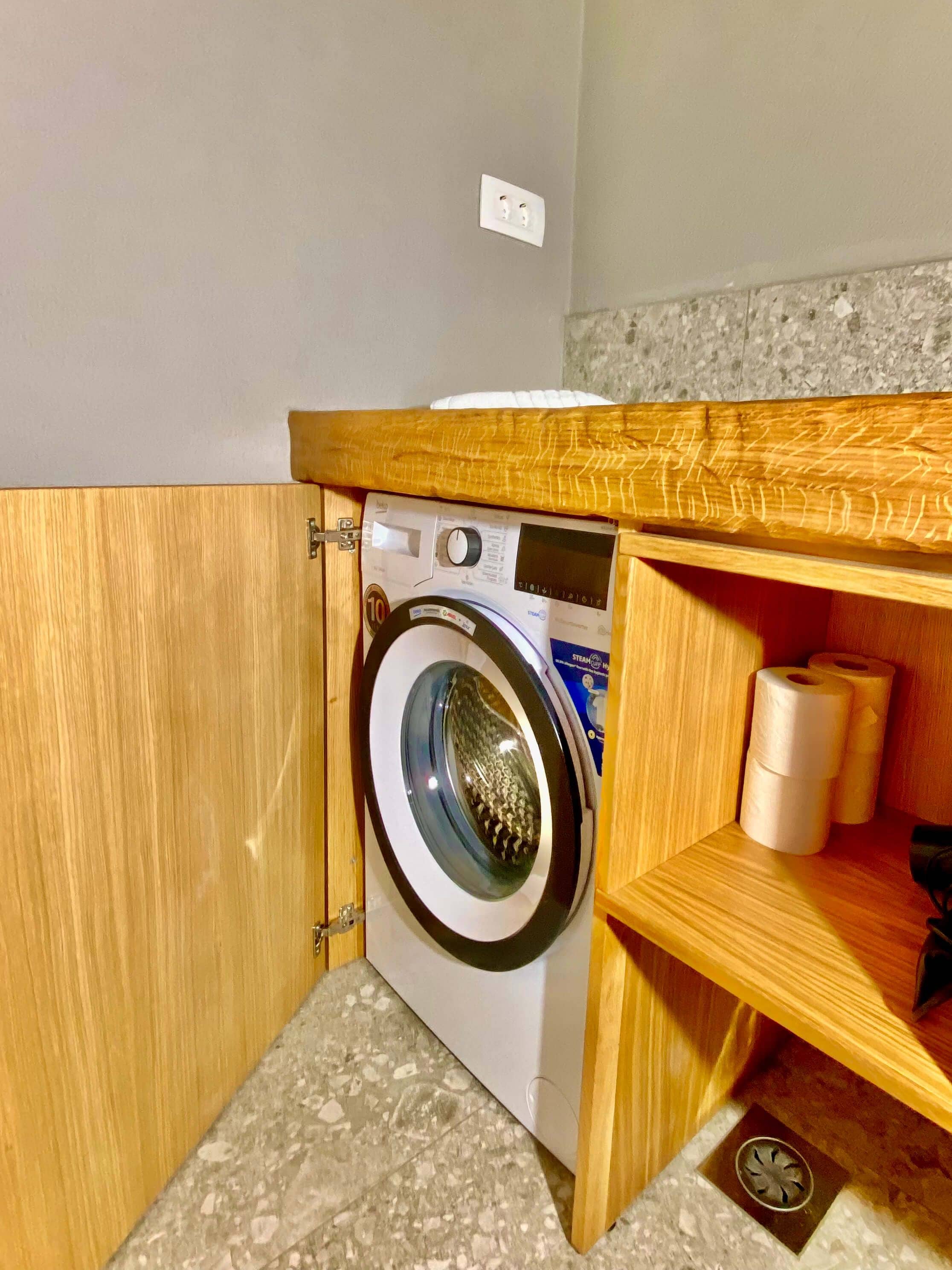 A compact laundry area with a washing machine, a shelf with toilet paper, and a wooden cabinet.