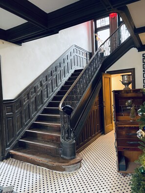 A grand staircase with ornate metal railings and a wooden finish, leading to a room with a patterned floor and a Christmas tree. - The Dery House  (Catasauqua)