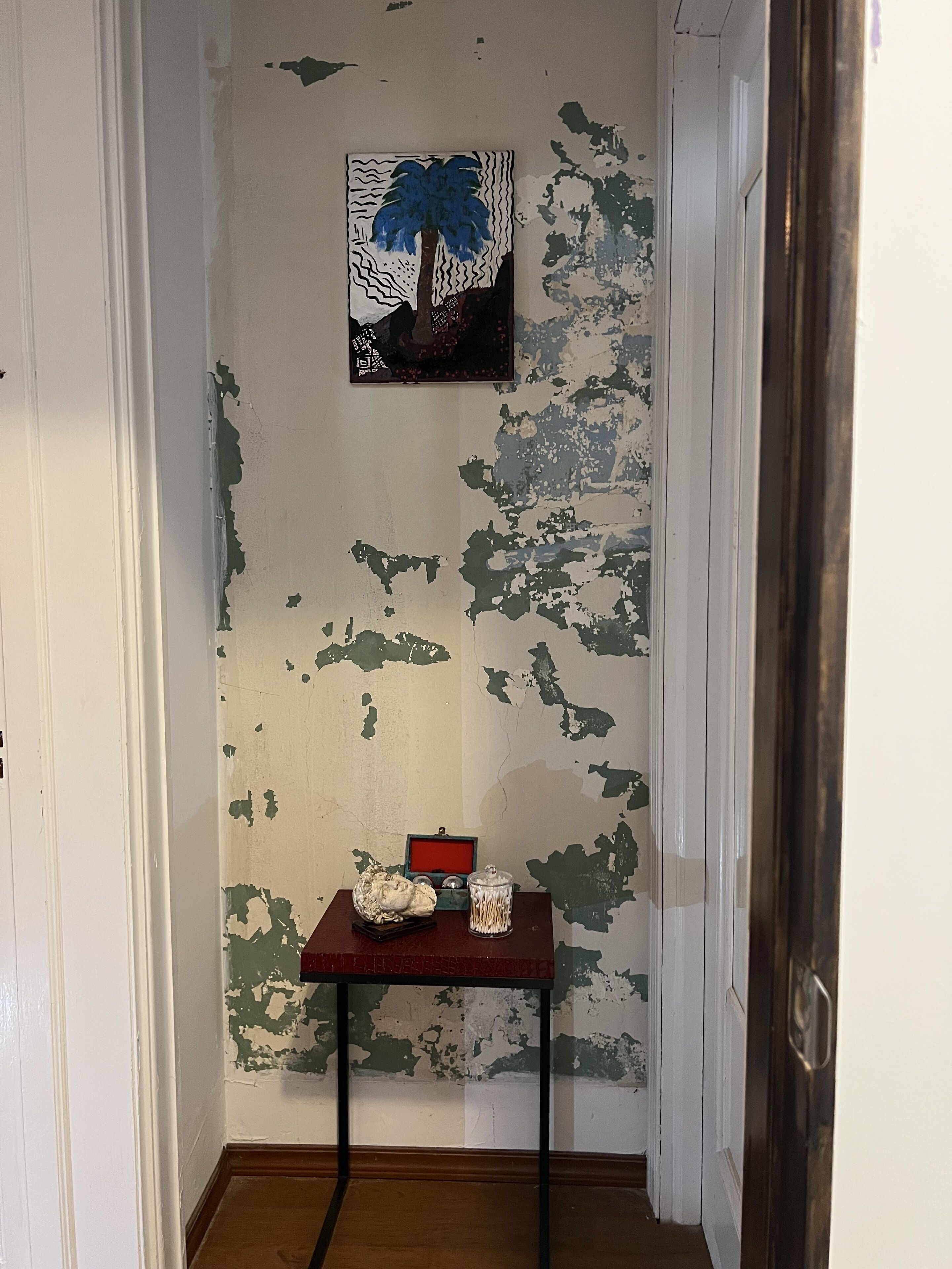 Um corredor com tinta descascando nas paredes, uma pequena mesa com um objeto decorativo e um pote de vidro, e um quadro com uma palmeira.