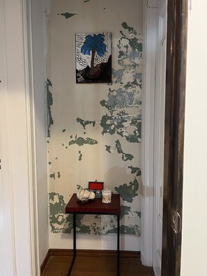 A hallway with peeling paint on the walls, a small table with a decorative item and a glass jar, and a framed picture of a palm tree. - The Dery House  (Catasauqua)
