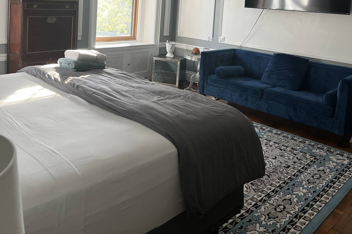 A bedroom with a bed, a blue sofa, a TV, a painting, and a window with blinds.