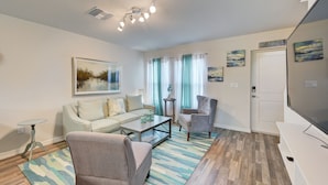 A living room with a sofa, armchairs, a coffee table, and a TV. There are curtains, a painting, and a sign on the wall. - Gulf Cost Home 5min to I-75 & Sunseekers Resort (Port Charlotte)