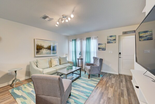 A living room with a sofa, armchairs, a coffee table, and a TV. There are curtains, a painting, and a sign on the wall.