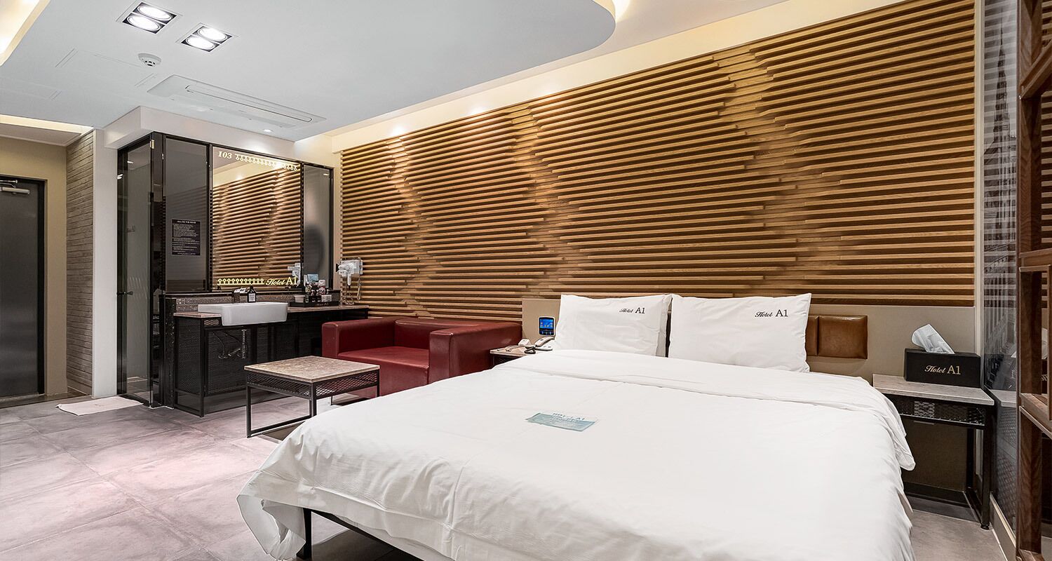 Ein modernes Hotelzimmer mit einem großen Bett, einem roten Sessel, einem Schreibtisch mit Spiegel und einer Wand aus Holzlamellen.
