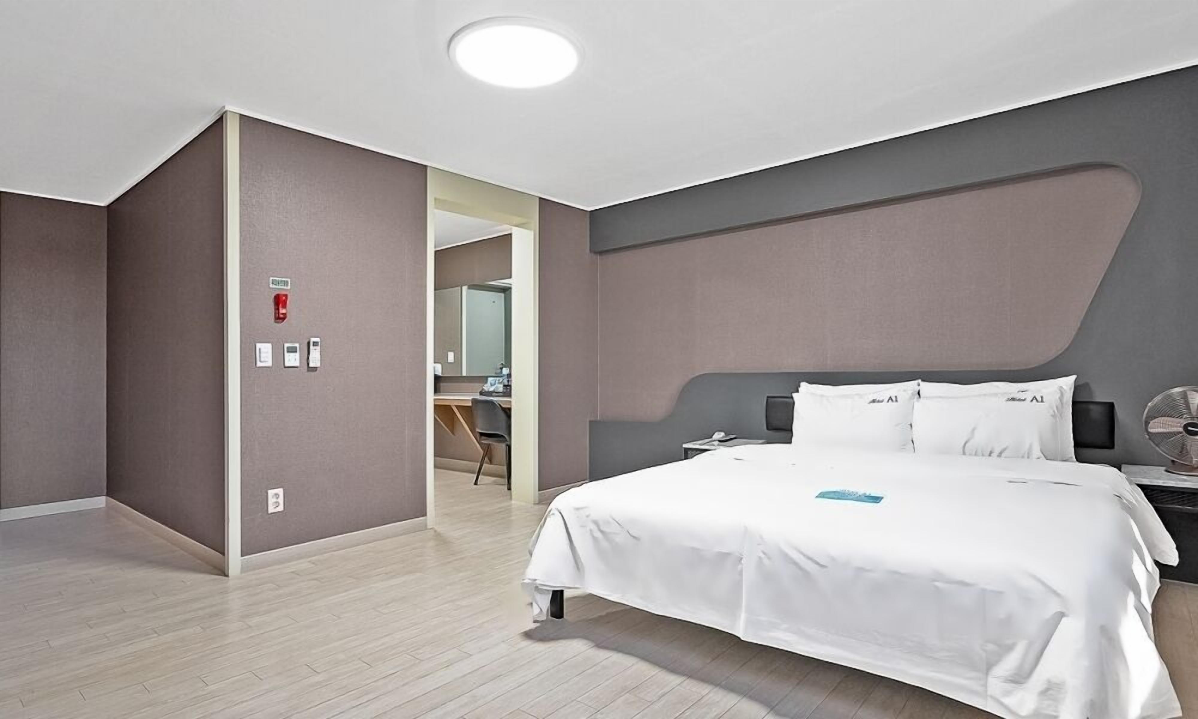 A modern hotel room with a large bed, a bedside table with a lamp, and a view of the hallway.