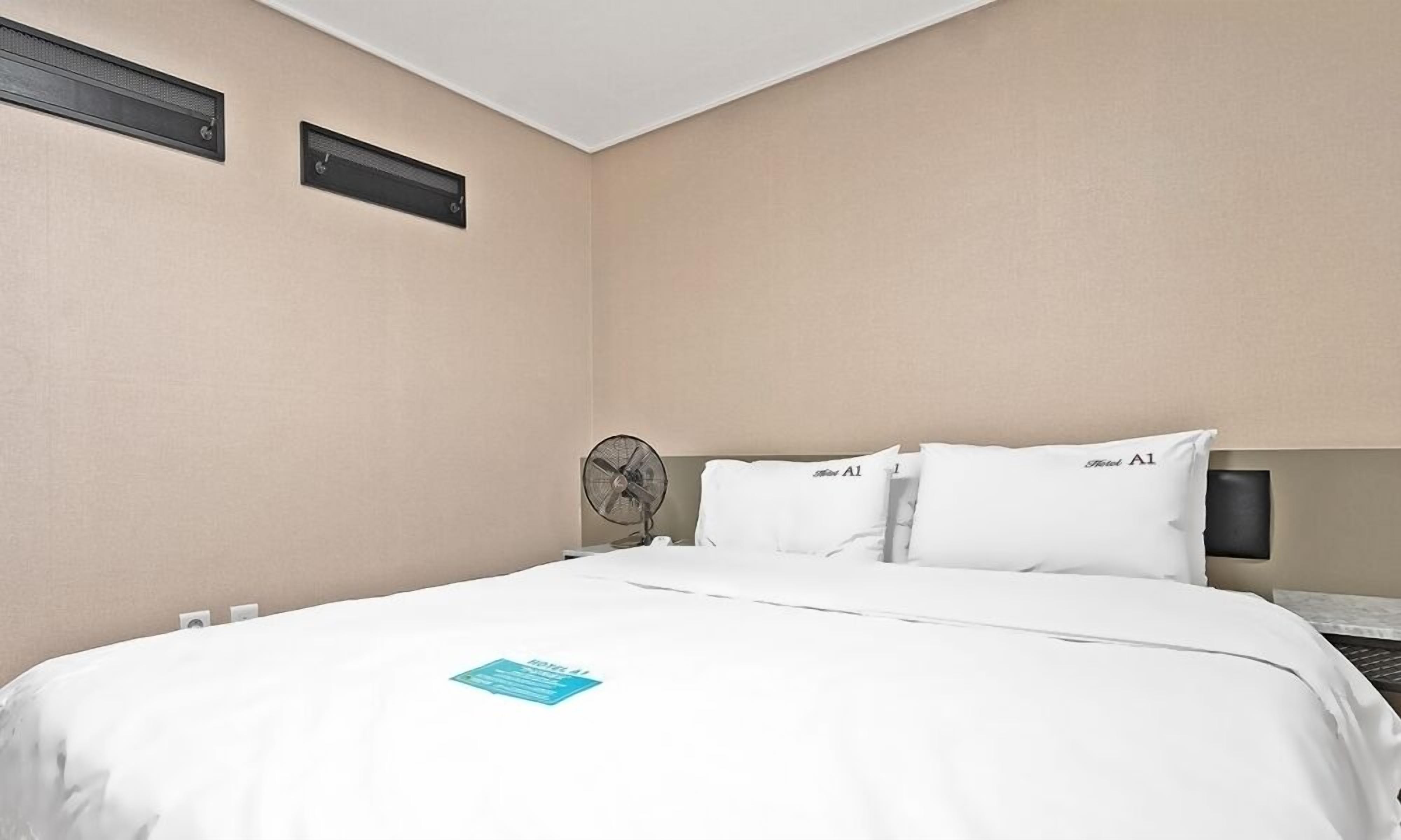 Ein Hotelzimmer mit einem Bett, zwei Kissen mit der Aufschrift 'Hotel A1', einem Nachttisch, einem Ventilator und zwei Luftauslässen an der Wand.