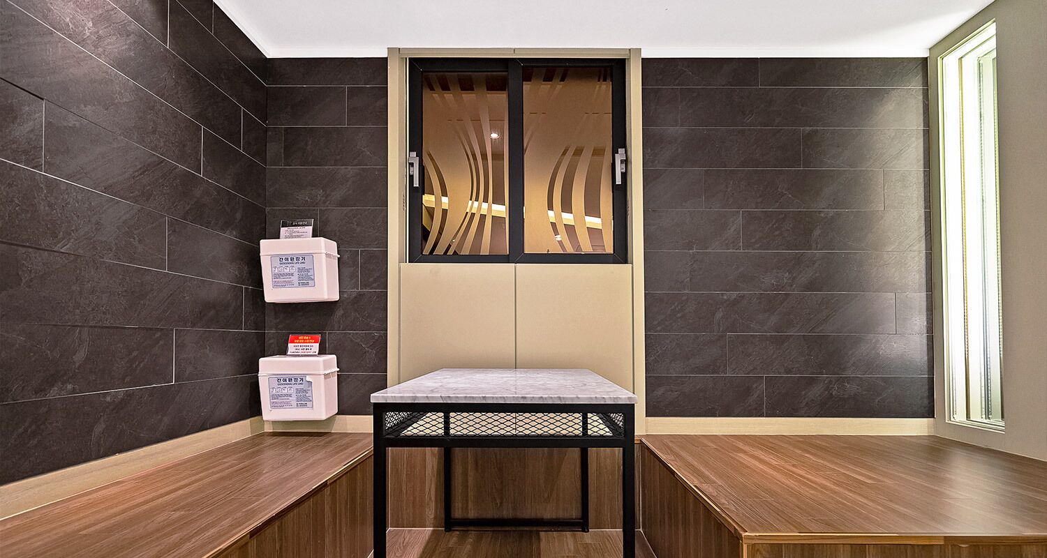A modern bathroom with dark tiled walls, a wooden bench, a small table, and a cabinet with toiletries.