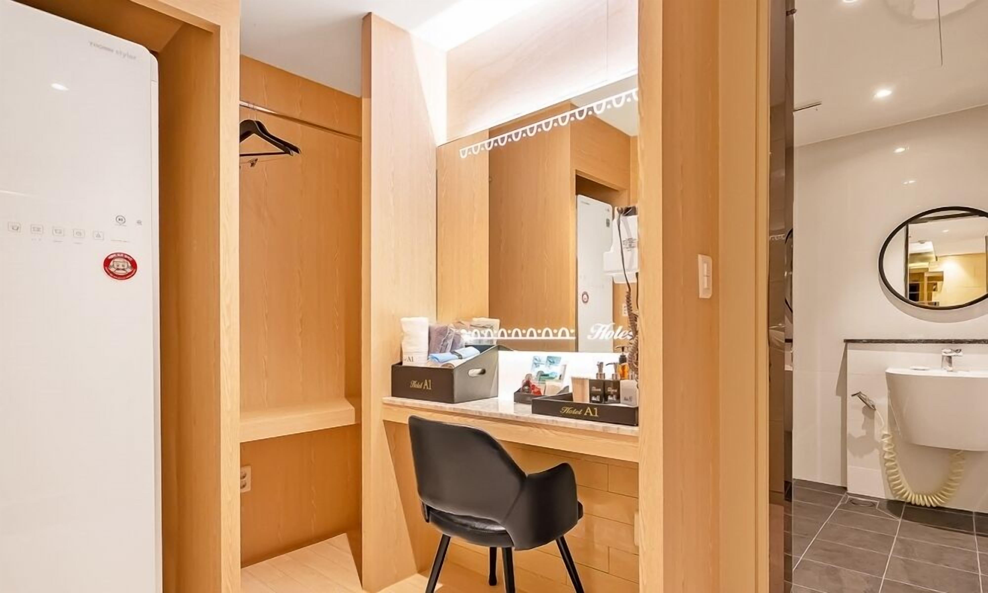 A hotel room with a wooden wardrobe, a black chair, a minibar, and a bathroom with a sink and mirror.