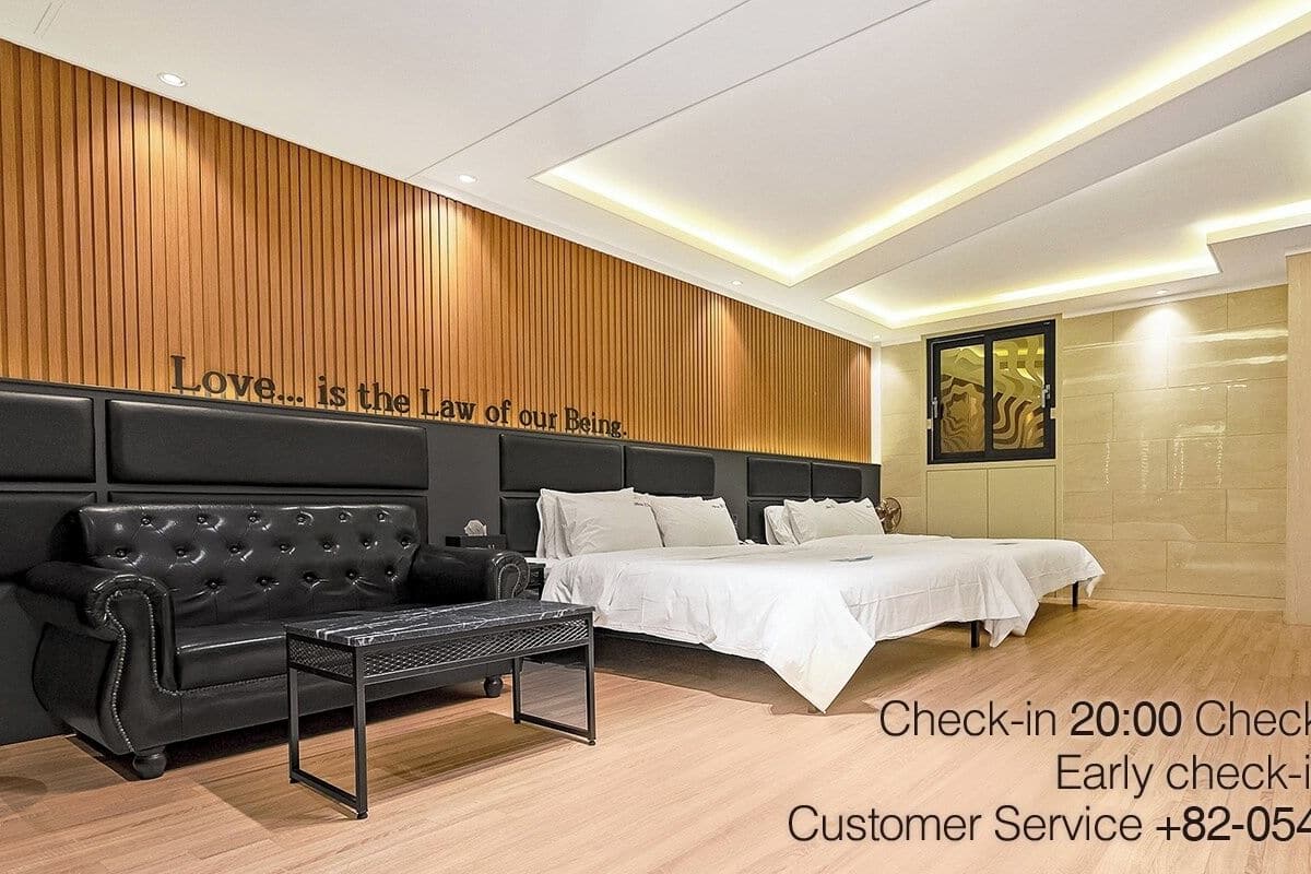 Habitación de hotel con sofá de cuero negro, cama con ropa blanca y pared de madera con una cita. La hora de llegada es a las 20:00 y la hora de salida a las 12:00.
