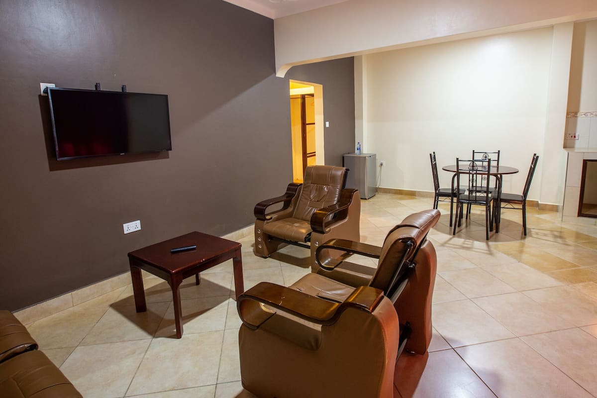 A living room with a flat-screen TV mounted on the wall, a brown leather sofa, a wooden coffee table, and a dining area with a small table and chairs.