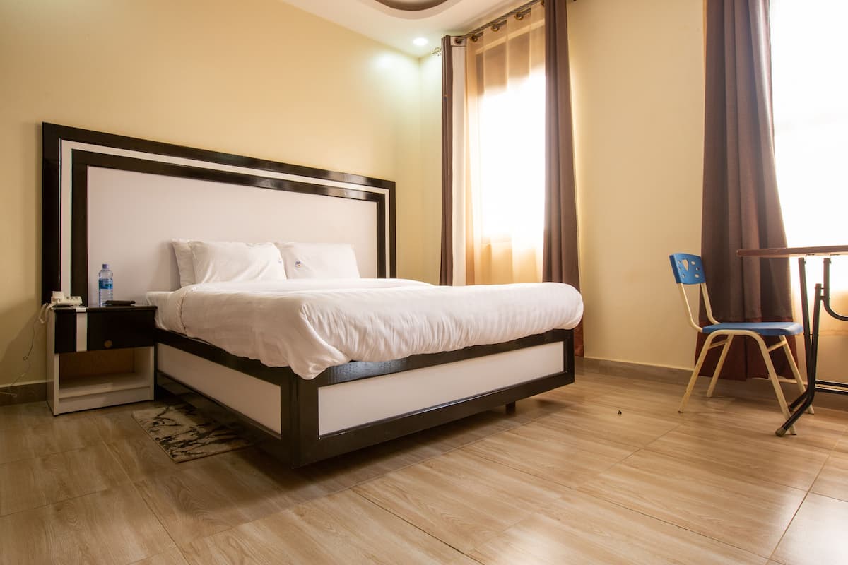 A neatly made bed with a wooden frame and white bedding, a small bedside table with a water bottle, and a chair in a room with wooden flooring and beige walls.