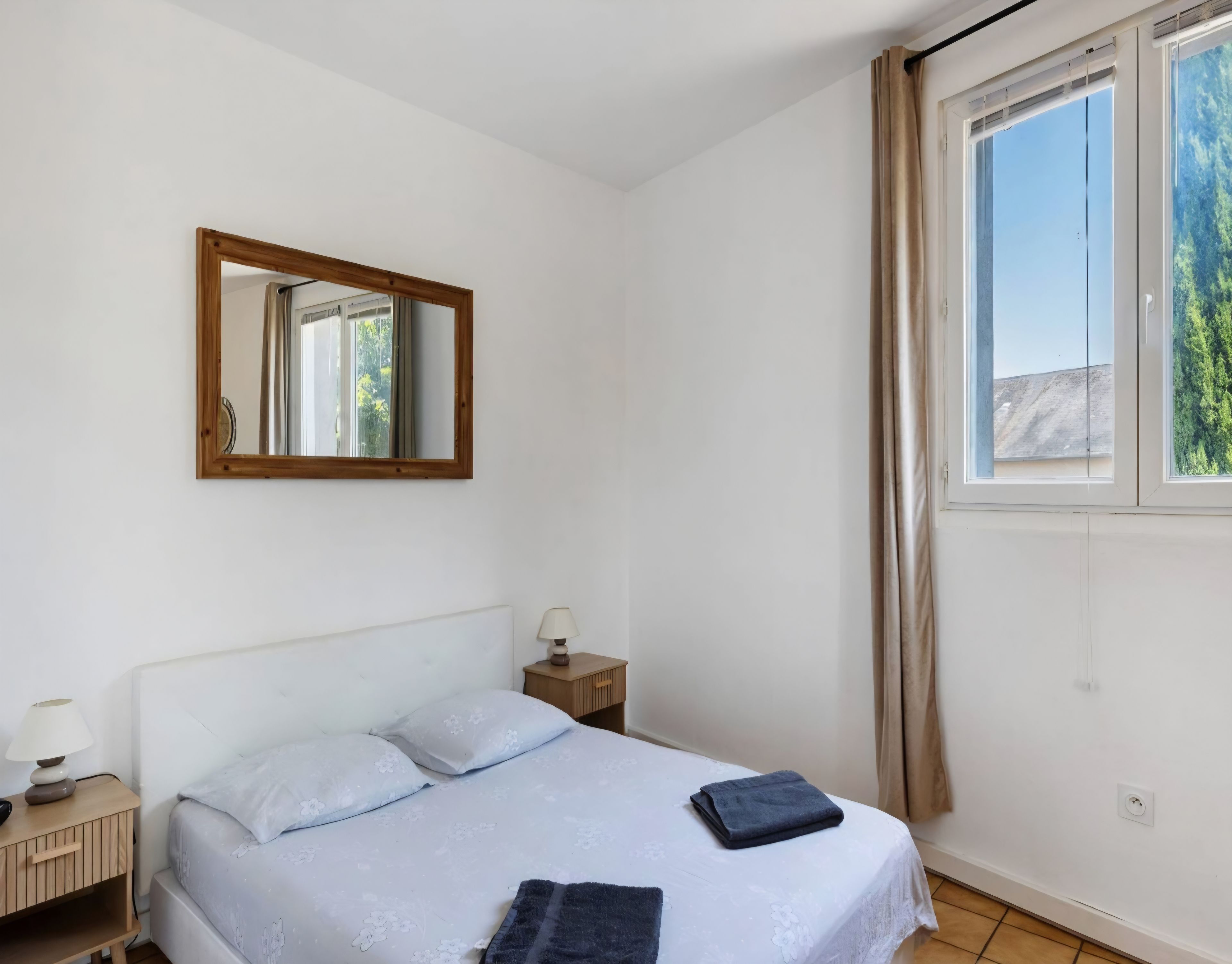 A bedroom with a bed, a nightstand, a lamp, a mirror, and a window with curtains.
