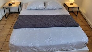 Una cama individual con colcha gris y ropa de cama floral, flanqueada por dos mesitas de noche con lámparas, en una habitación con suelo de baldosas y un cuadro colgado en la pared.