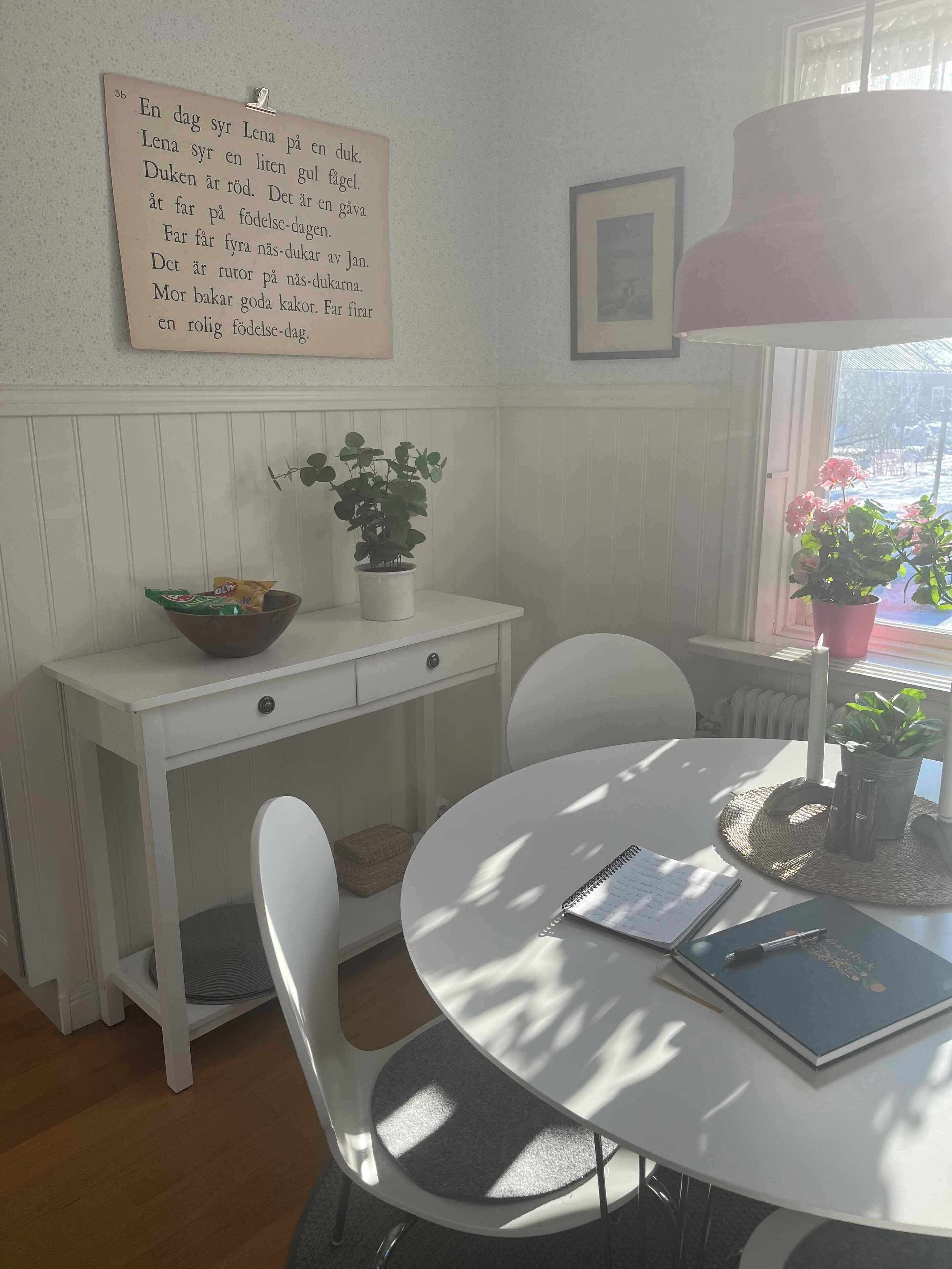 A room with a white desk, a round table, and chairs. There is a potted plant on the desk, a framed picture on the wall, and a window with a view of the outdoors.