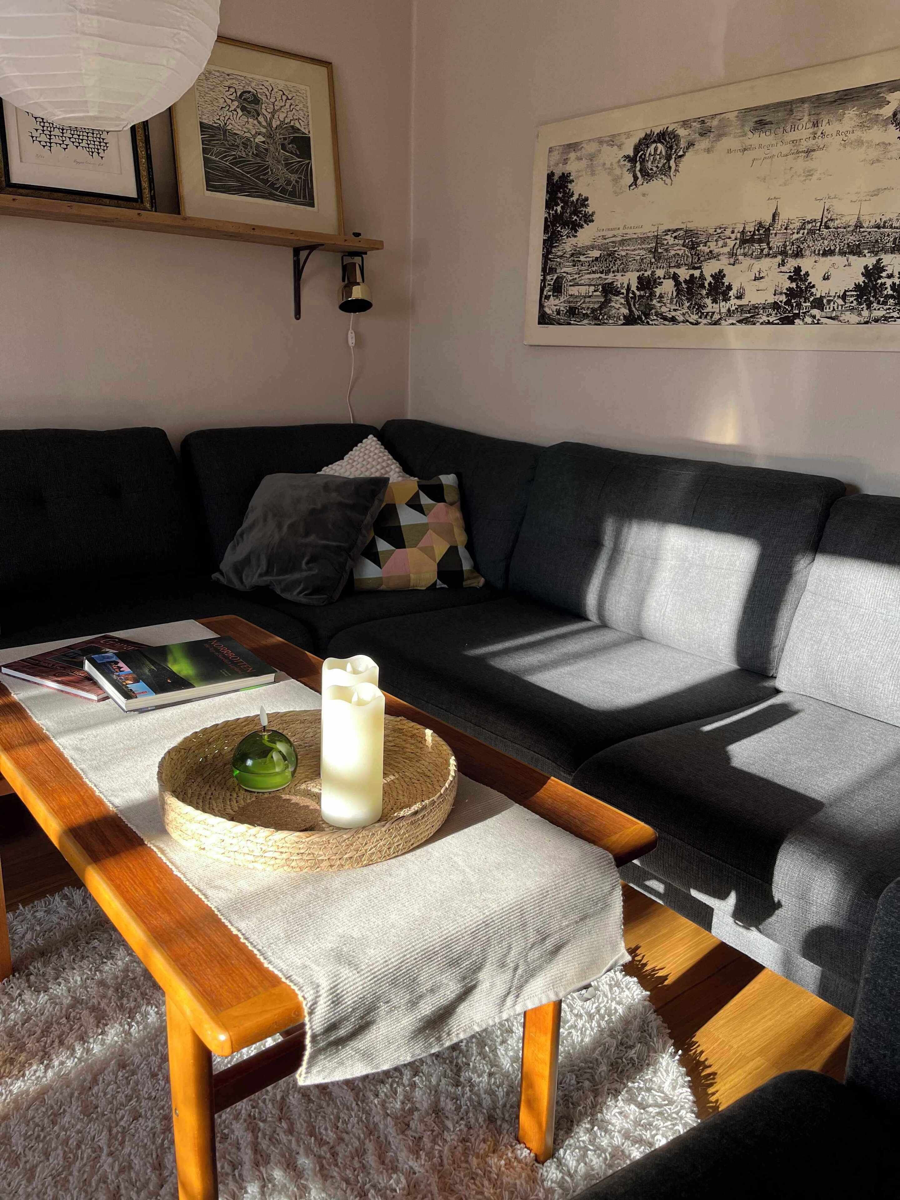 Un salon moderne avec un canapé gris, une table basse en bois sur laquelle sont posés une bougie et un livre, et une œuvre d’art encadrée représentant un paysage urbain accrochée au mur.