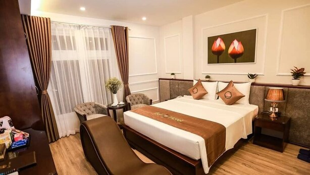 A hotel room with a large bed, a sitting area, a desk, and a window with curtains. - Sen Hotel by Bay Luxury (Hanoi)