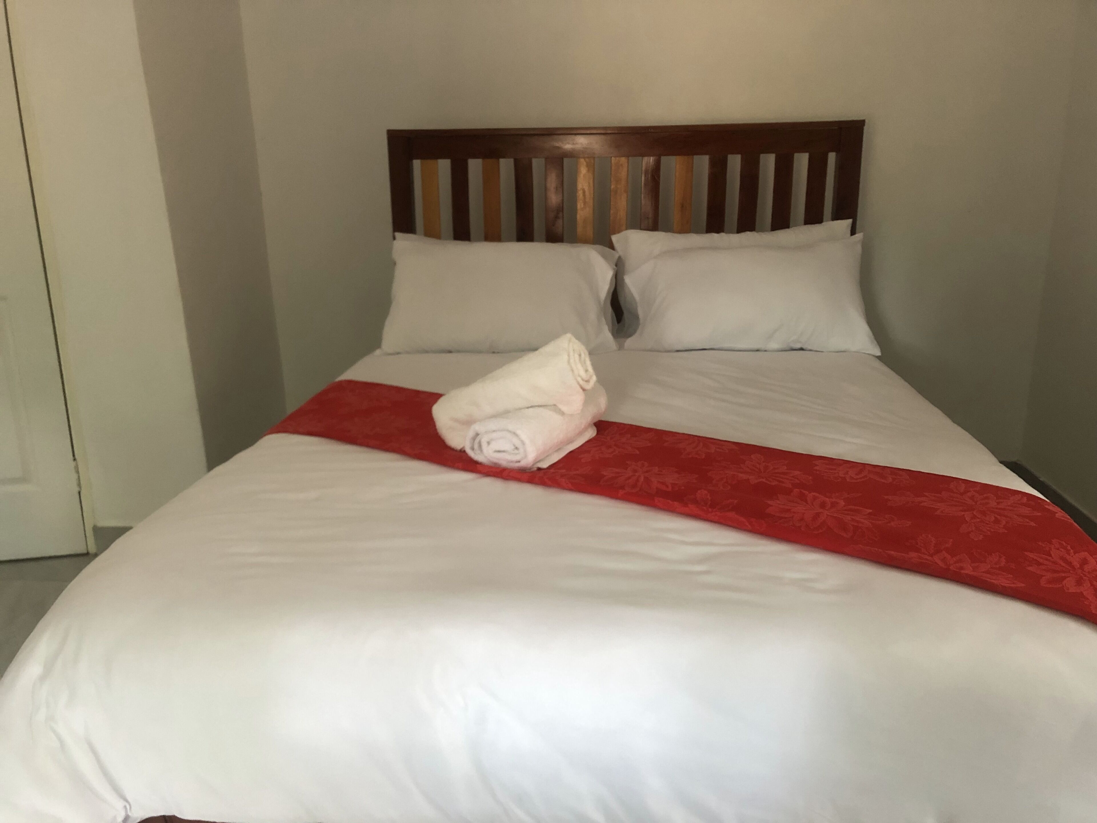 Una cama bien hecha con sábanas blancas y un detalle rojo, un cabecero de madera y una puerta blanca a la izquierda.