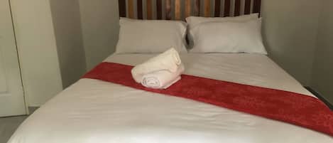 A neatly made bed with white linens and a red accent, a wooden headboard, and a white door to the left.