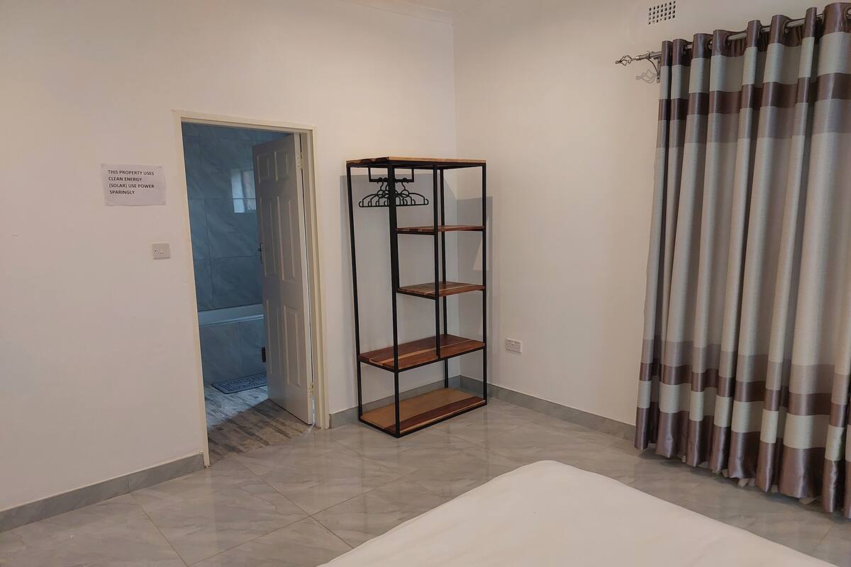 Ein Zimmer mit Fliesenboden, einem weißen Bett, einem Metallregal, einer Tür mit einem Schild und einem Vorhang.