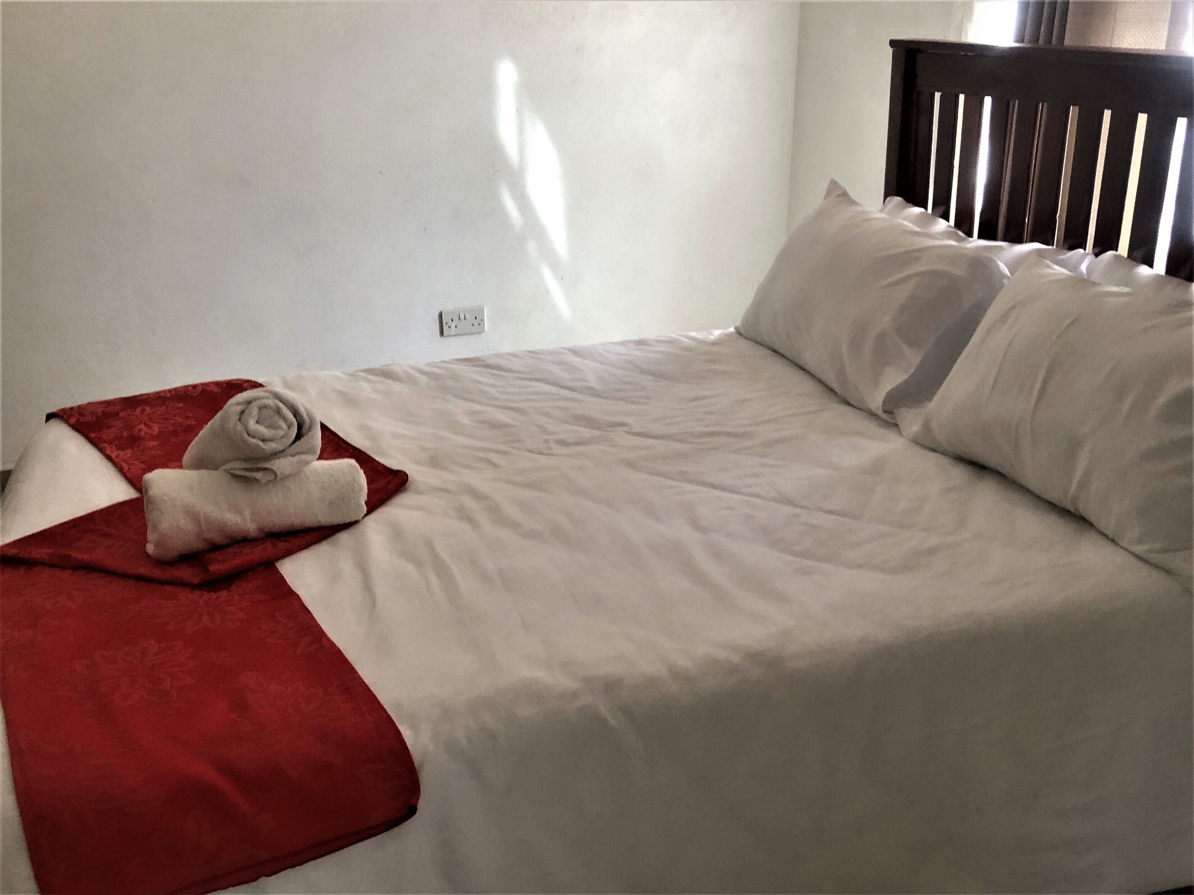 Ein ordentlich bezogenes Bett mit weißen Leinentüchern und einer roten Decke, ein hölzerner Kopfteil und eine Steckdose an der Wand.