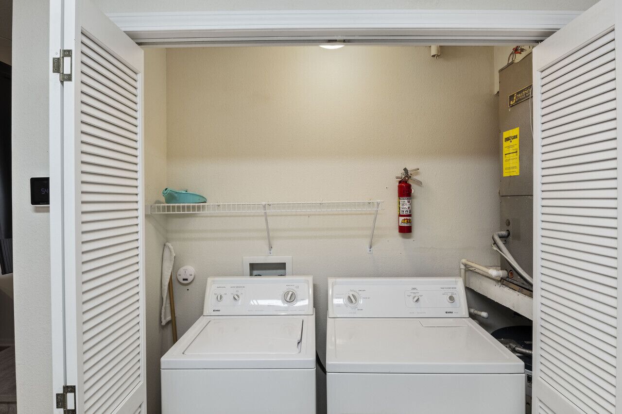 A laundry room with two white front-loading washers and dryers, a fire extinguisher on the wall, and a shelf above the dryers.
