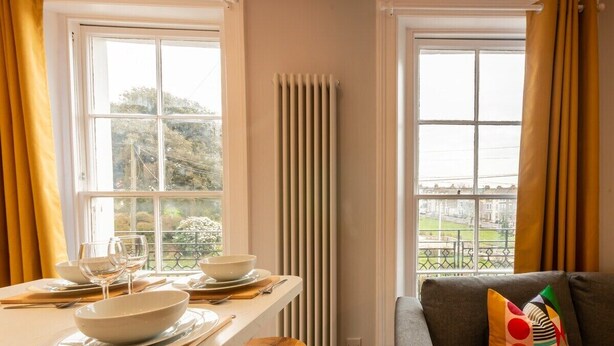 Een zithoek met een eettafel gedekt voor twee personen, een radiator en een bank met een kleurrijk kussen.