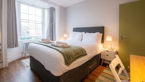 Een net opgemaakt bed met witte lakens en een groene plaid, een nachtkastje met een lamp, een raam met gordijnen, en een groene deur.