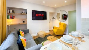 Een moderne woonkamer met een grijze bank, een gele fauteuil, een flatscreen televisie, een boekenkast en een eethoek met borden en glazen.