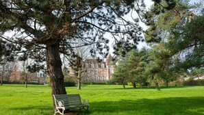A park with a bench under a large tree, a grassy field, and a building in the background. - Un Appartement Dans un Château (Deauville)