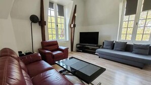A modern living room with a leather sofa, a grey couch, a flat-screen TV, and a large window. - Un Appartement Dans un Château (Deauville)