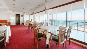 A dining area with tables and chairs, large windows with a view of the sea, and a red carpeted floor. - Miletus Otel (Sile)