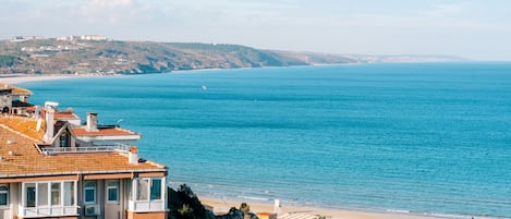 Une vue côtière avec des bâtiments surplombant la mer, une plage et des collines à l’horizon.
