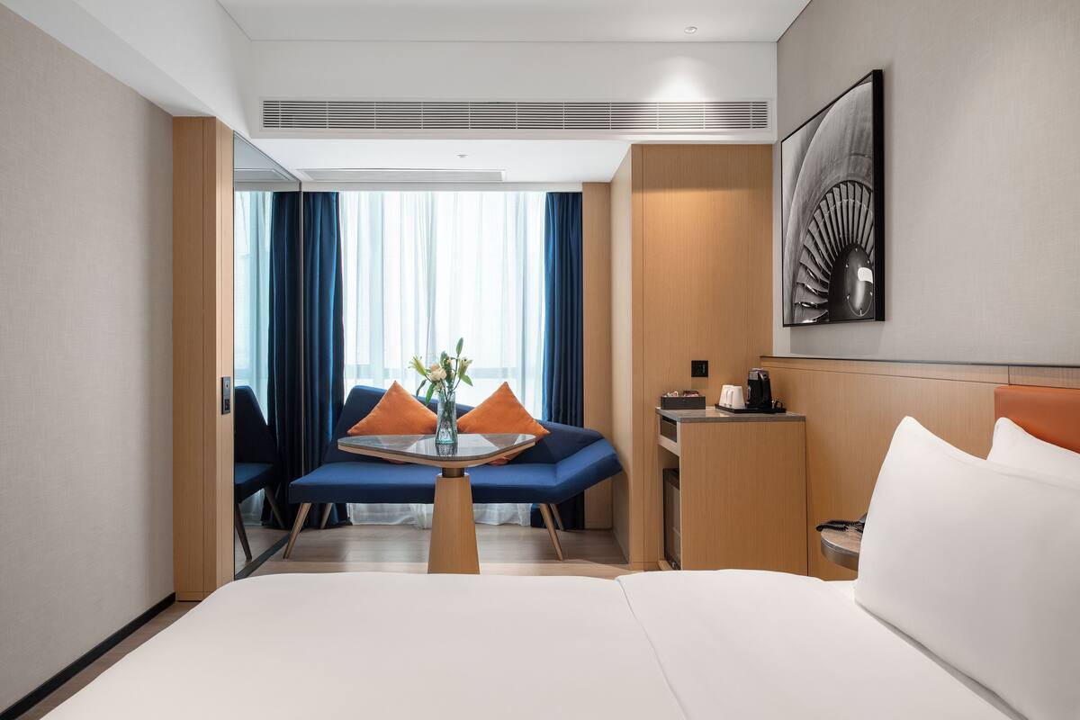 Ein Hotelzimmer mit Bett, kleinem Tisch mit Vase, einer Couch mit Kissen, einem Schreibtisch mit Kaffeemaschine und einem großen Fenster mit Vorhängen.