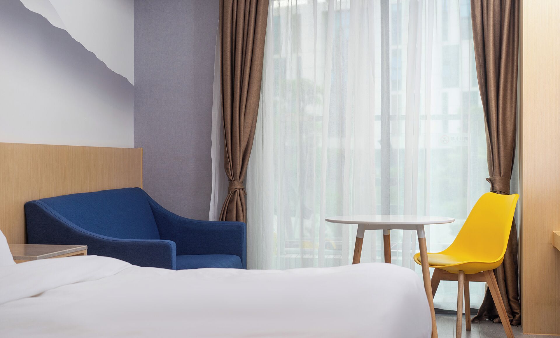 A hotel room with a bed, a blue armchair, a yellow chair, a small round table, and sheer curtains.