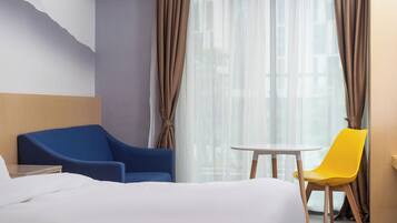 A hotel room with a bed, a blue armchair, a yellow chair, a small round table, and sheer curtains.