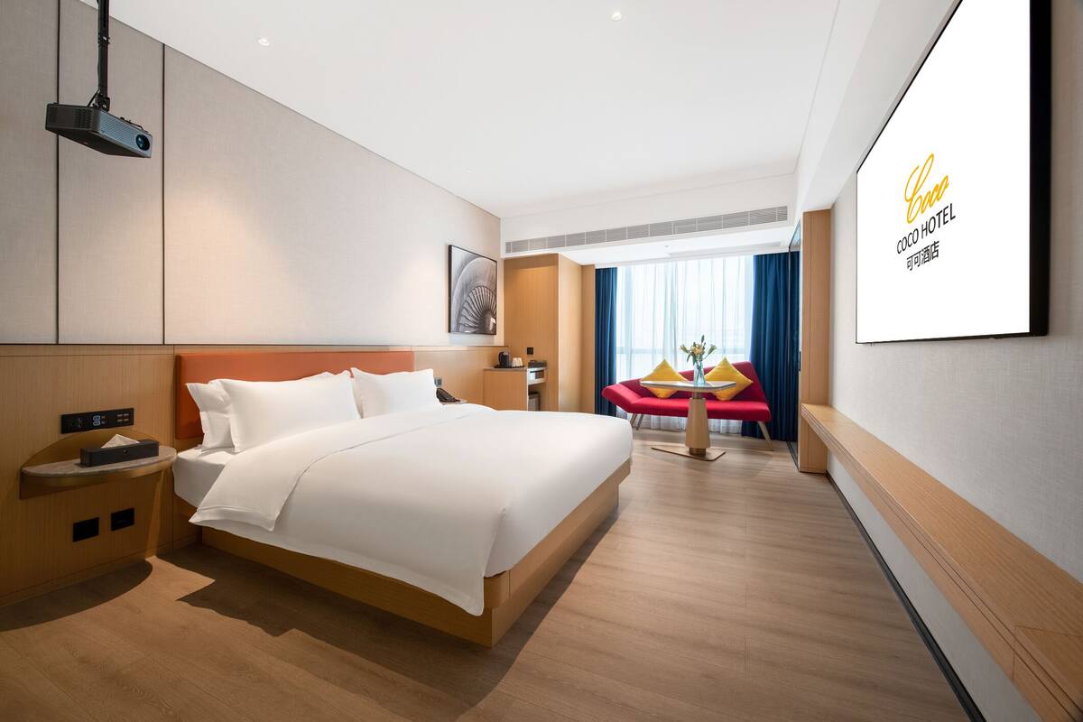 Ein modernes Hotelzimmer mit einem großen Bett, einem Nachttisch, einem Projektor und einem großen Bildschirm, der den Namen des Hotels zeigt.