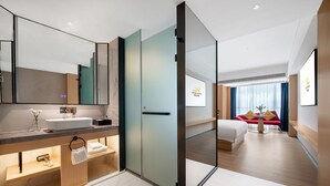 Ein modernes Hotelzimmer mit einem großen Spiegel, einem Waschbecken, einem Nachttisch, einem Bett und einer Schiebetür zu einem weiteren Zimmer.