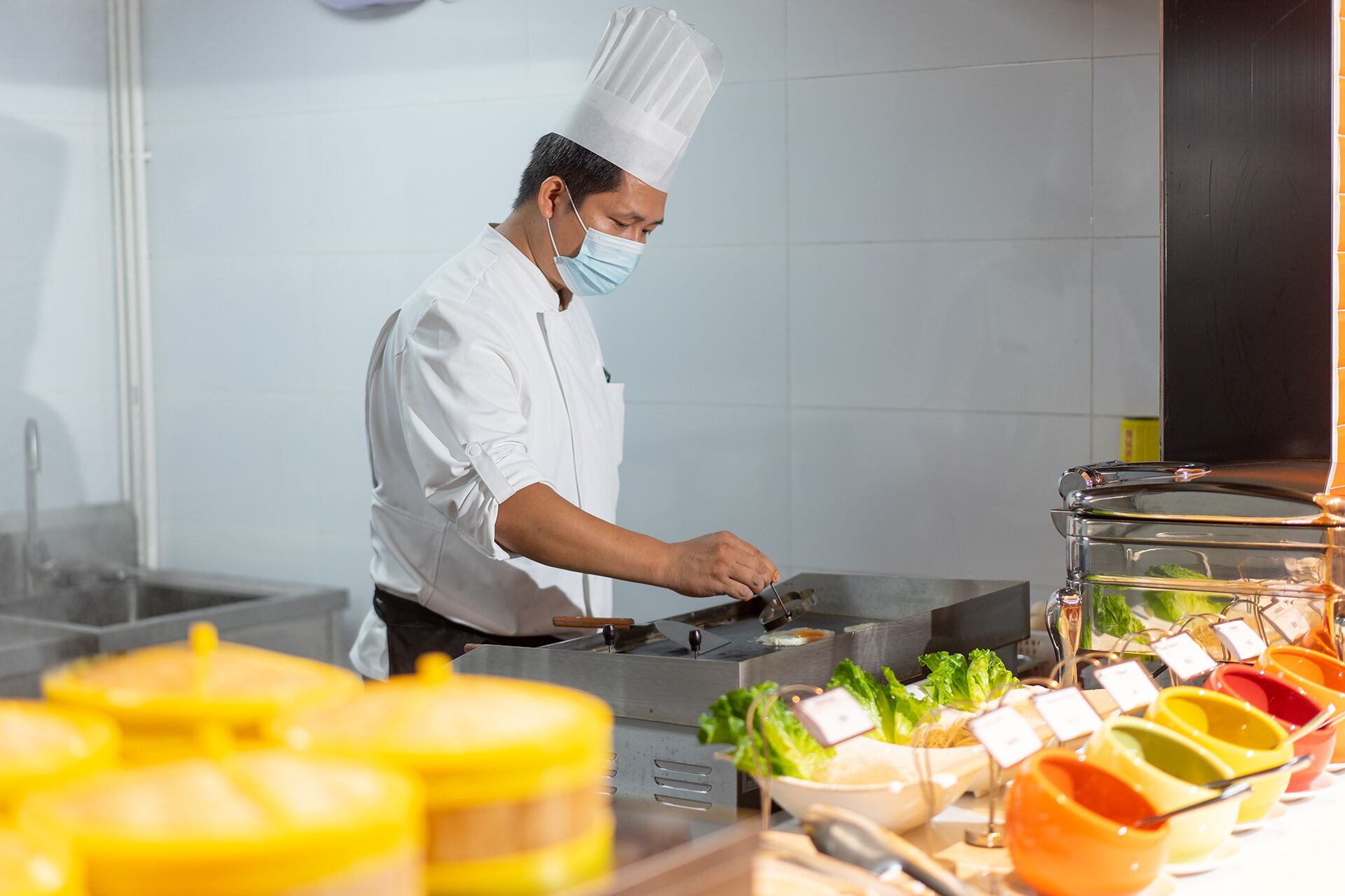 A chef in a white uniform and hat preparing food in a kitchen with a buffet of colorful dishes.