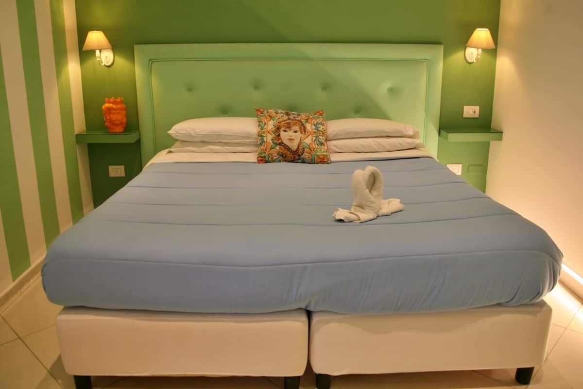Een bed met een blauwe matras, witte kussens en een sierkussen met een bloemendessin. Twee nachtkastjes met lampen, en een kleine oranje vaas op één nachtkastje.