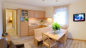 A modern kitchen with wooden cabinets, a dining area with a table and chairs, a small sofa, and a TV mounted on the wall.