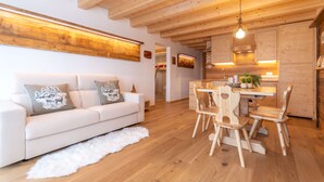 A modern interior with a wooden ceiling, a white sofa with decorative pillows, a dining area with wooden chairs, and a kitchen with wooden cabinets and a hanging light fixture. - Chalet Laura (Alleghe)