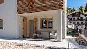 A modern building with a covered patio area, a small table with chairs, and a view of a hillside with other buildings. - Casa Carducci (Alleghe)